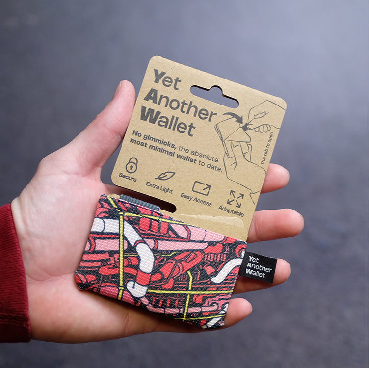 Una mano sostiene una cartera YAW con estampado rojo y amarillo junto a su envoltorio de papel kraft